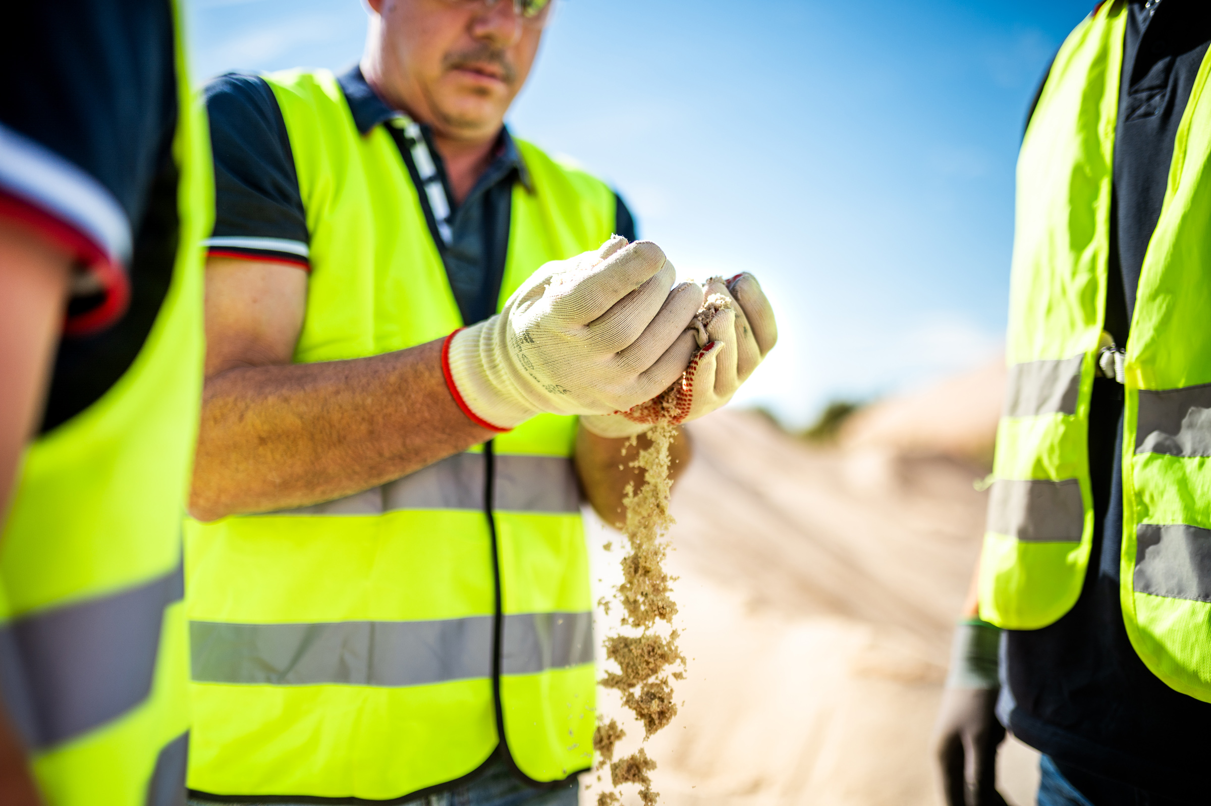 Ein Mitarbeiter des Quarzsandwerkes Wellmersdorf lässt Quarzsand durch seine Hände rieseln. Im Hintergrund ist ein großer Sandhügel zu erkennen.