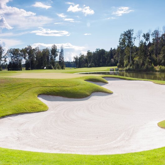 Well-maintained golf course featuring a large white sand bunker beside a green lawn and a pond in the background.