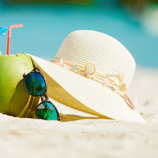 Green coconut with straw and cocktail umbrella, next to a sun hat and sunglasses on white beach sand.
