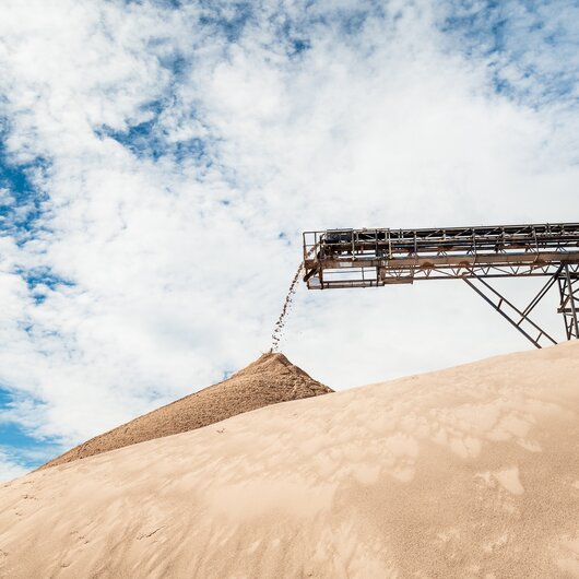 Conveyor belt pouring sand onto a large pile under a partly cloudy blue sky.