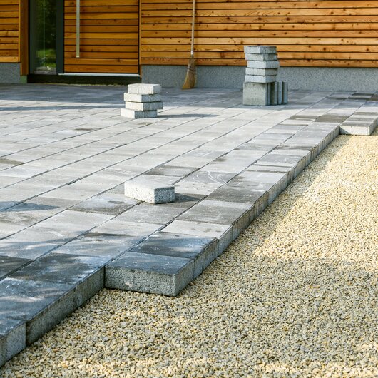 Partially laid gray concrete slabs forming a new terrace beside a wooden house; gravel visible on the side.