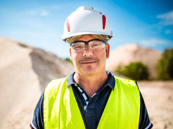 A worker wearing a helmet, goggles, and a yellow vest stands in front of sand mounds, facing the camera.
