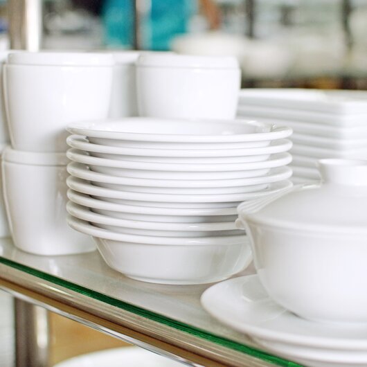 Stacked white porcelain dishes with bowls, cups, and plates on a glass shelf in a kitchen or cafeteria.