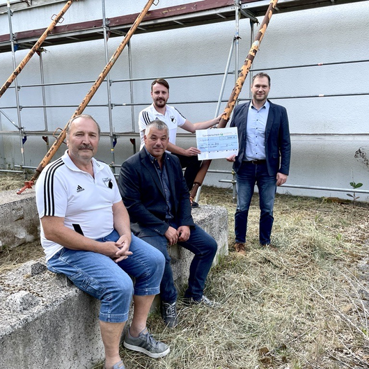 Four men sit and stand in front of a construction site; one symbolically presents a large donation check.