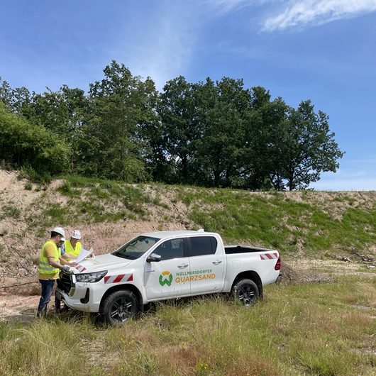 Two people in safety gear stand beside a white company pickup labeled “Wellmersdorfer Quarzsand,” reviewing documents at a sand site.