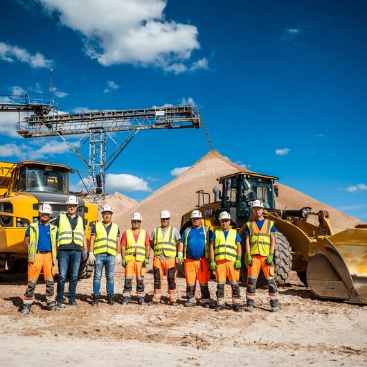 Group of workers in safety gear standing in front of sand piles, construction vehicles, and a conveyor belt.