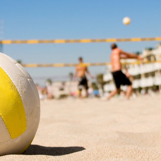 A volleyball lies on the sandy ground while people play beach volleyball in the sunny background.