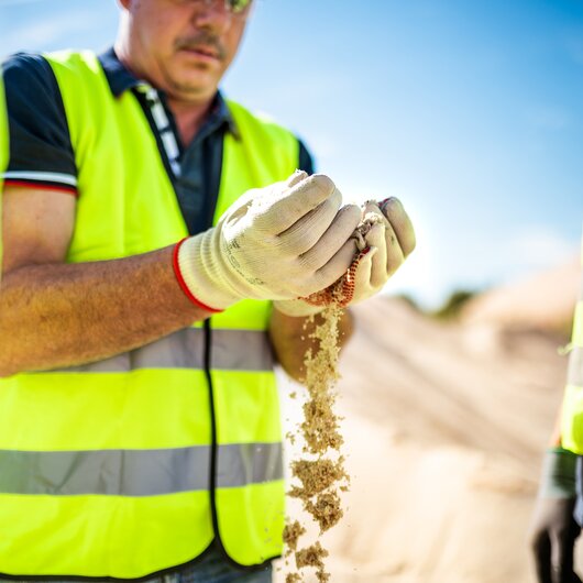 A worker in a safety vest and gloves lets sand run through his hands while inspecting the material.