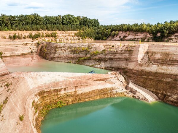 Panorama of a flooded quartz sand excavation site with terraced sandstone walls and turquoise water, surrounded by dense vegetation.